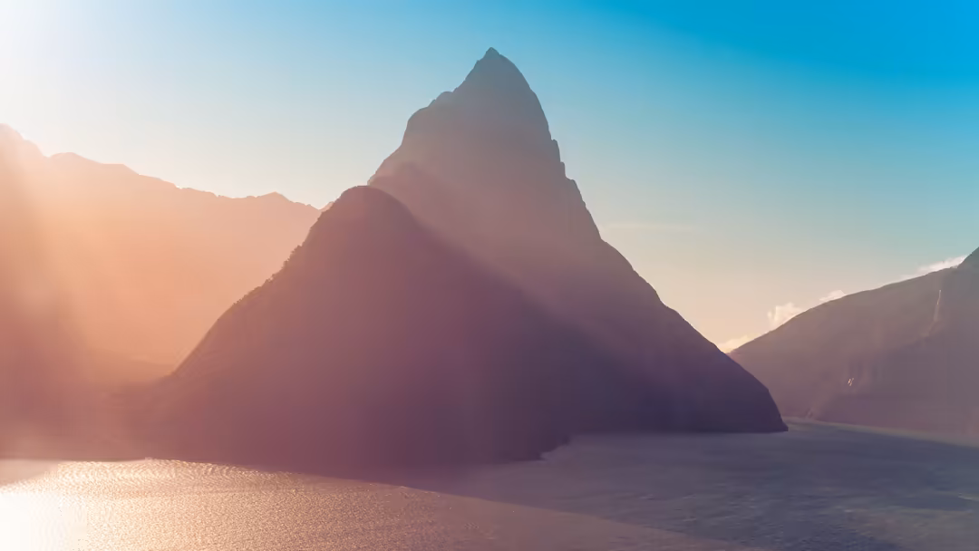 Dramatic mountain peak rises from calm alpine lake at sunset, bathed in warm golden light with misty atmosphere and surroundi