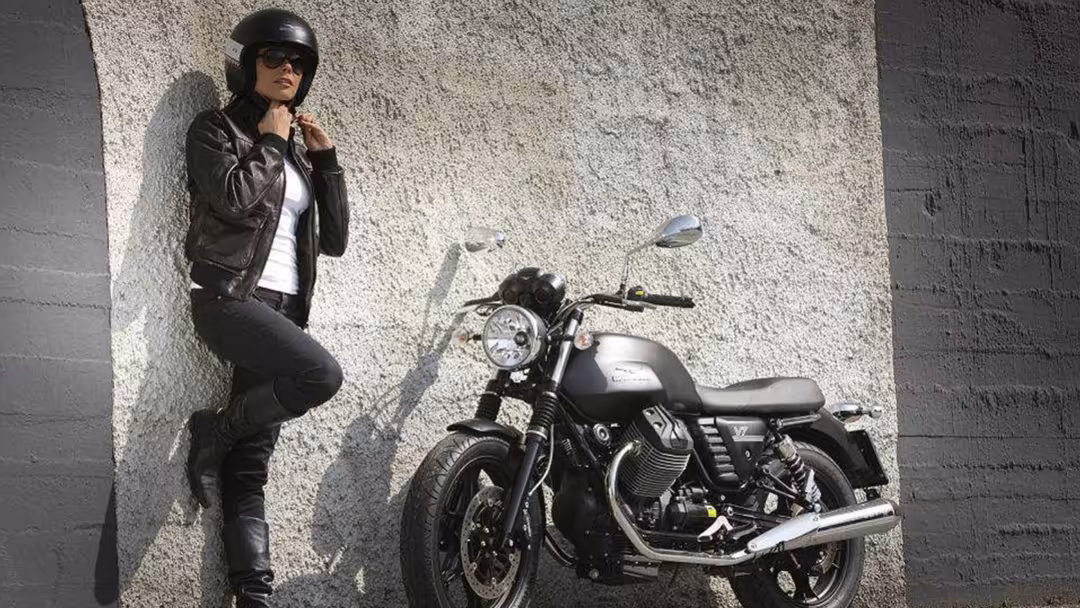 Stylish rider in black leather jacket and helmet standing against concrete wall next to classic motorcycle