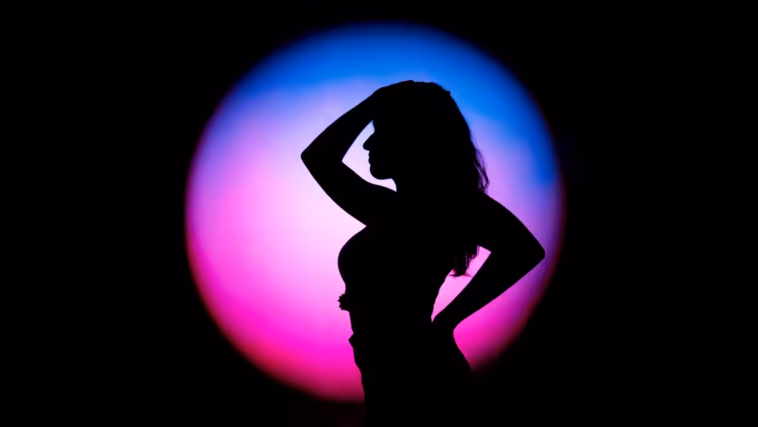 Black silhouette of a woman with hand to head against a vibrant neon pink and blue circular glow on dark background