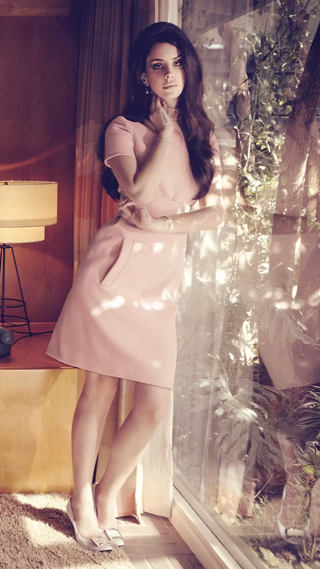 Woman in pink dress standing by illuminated plants and warm lamp lighting, vintage aesthetic with moody atmospheric ambiance