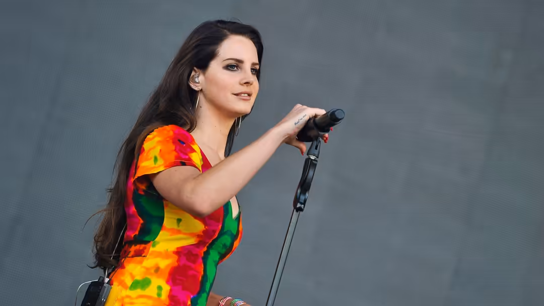 Singer performing on stage wearing vibrant multicolored outfit, holding microphone against gray background with focused expre