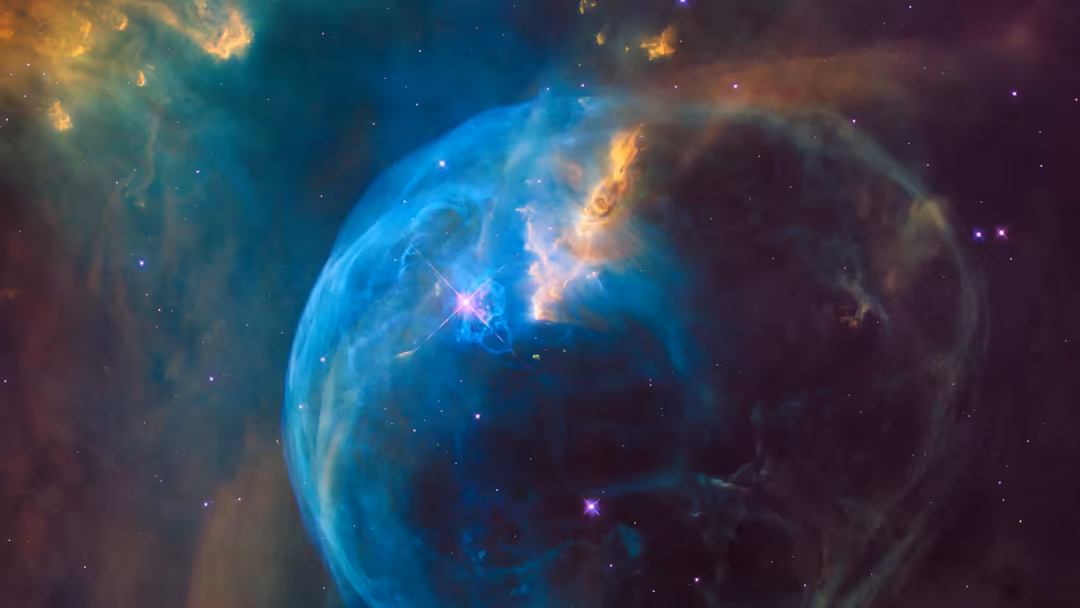 Brilliant blue bubble nebula with glowing orange gas wisps and bright stars scattered throughout the cosmic dust cloud