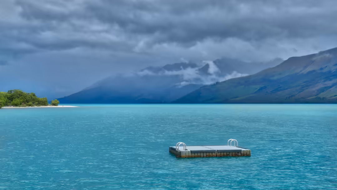 Pristine turquoise lake with wooden dock, green trees on white sand beach, and dramatic cloud-covered mountain peaks in backg