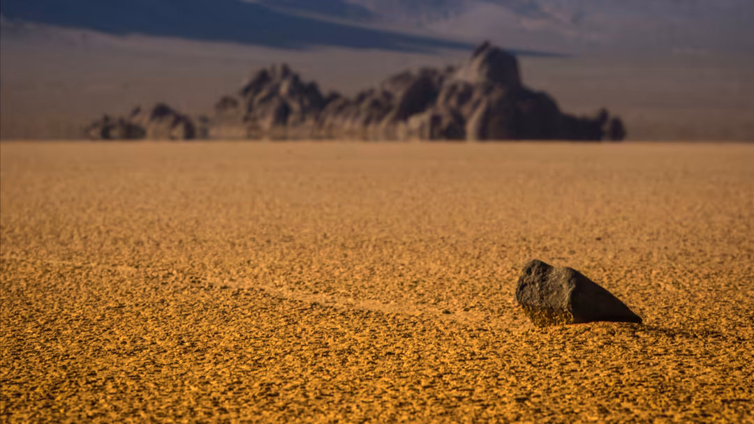 A solitary rock sits on vast golden desert sand with jagged rock formations visible in the hazy distance under a dusty sky