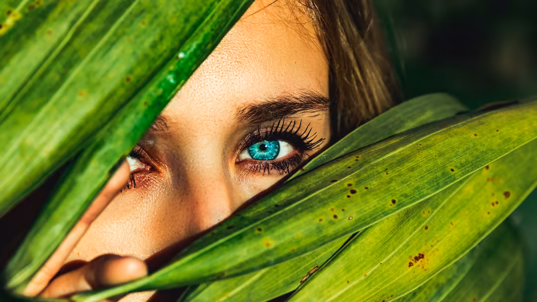 Woman with striking blue eyes peeking through vibrant green tropical leaves in natural sunlight