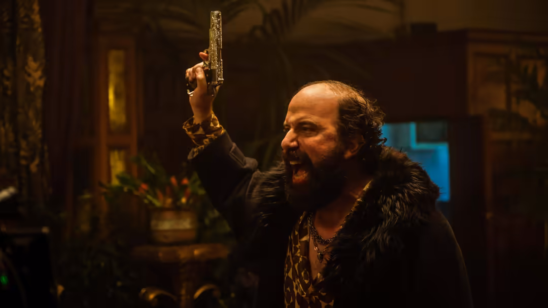 Bearded man with intense expression holds an ornate revolver aloft in a dark, moody interior with warm and cool lighting.