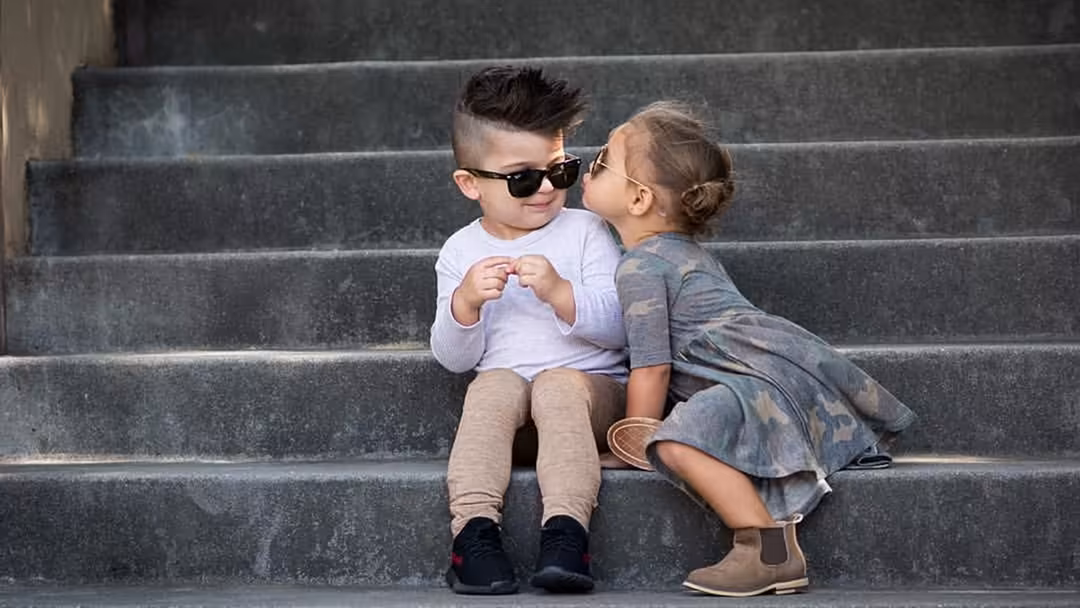 Two young children sitting together on concrete steps, one wearing sunglasses and white shirt, the other in patterned dress,