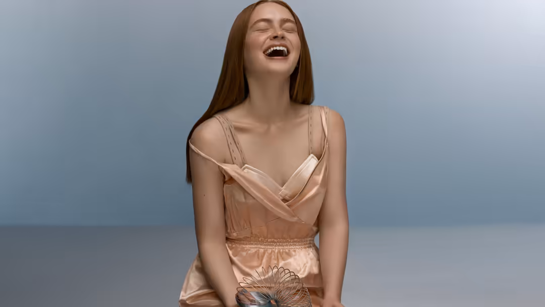 Young woman with long brown hair wearing a peach-colored wrap dress, smiling joyfully against a soft blue-gray background wit