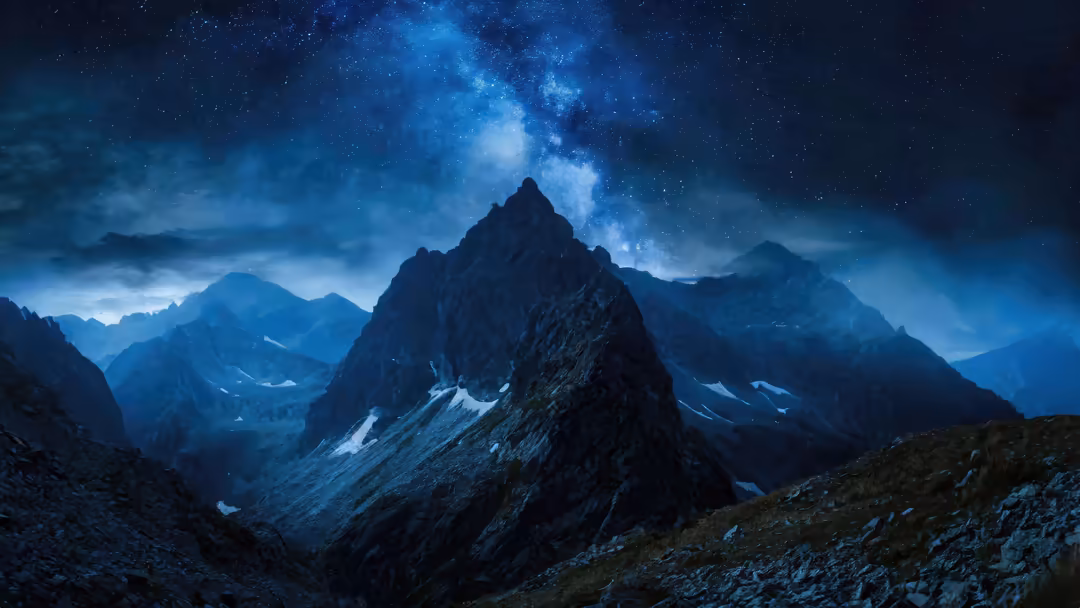Dramatic mountain landscape with snow-capped peaks silhouetted against a vibrant blue starry night sky and glowing milky way