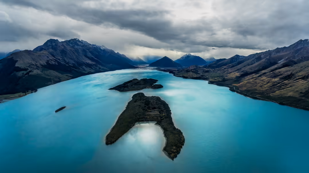 Aerial view of a stunning turquoise glacial lake surrounded by dramatic mountain peaks and dark volcanic islands under cloudy