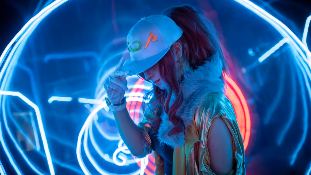 Figure wearing K/DA Akali baseball cap surrounded by glowing neon blue and pink lines in cyberpunk aesthetic setting with dra
