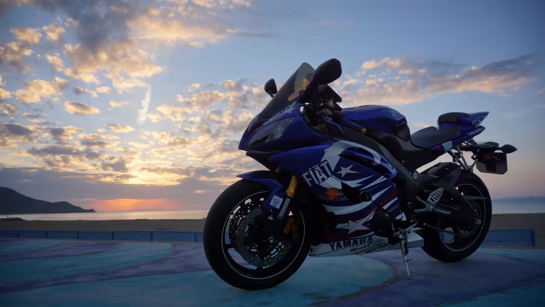 Blue sport motorcycle parked on coastal platform overlooking sunset ocean with golden clouds and distant mountains in backgro