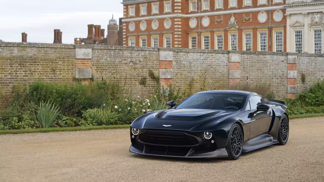 Sleek black Aston Martin Victor sports car parked on gravel driveway with historic brick palace and garden wall behind