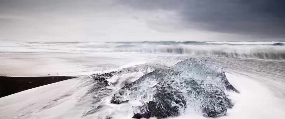 Dramatic coastal scene with massive ice formations on black sand beach, powerful waves crashing in background under stormy sk