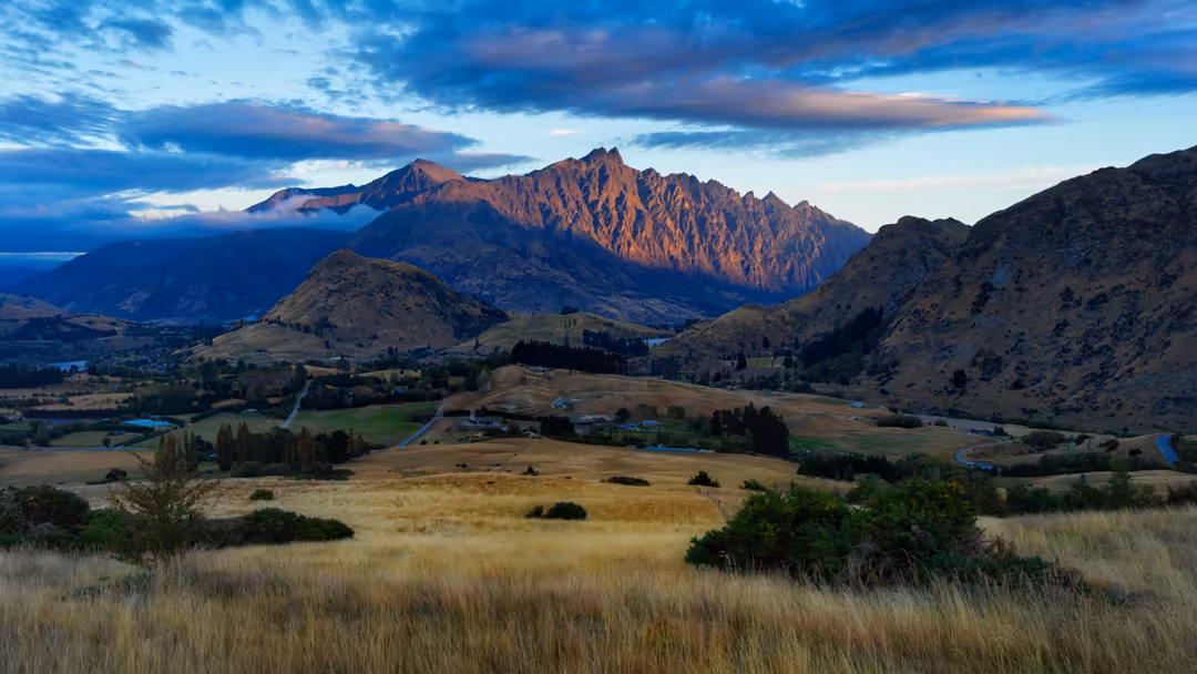 Dramatic alpine valley with rugged mountain peaks, golden grassland, winding river, and dramatic blue sky with clouds at suns