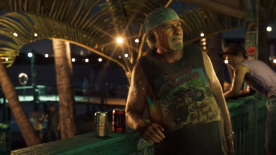 Bearded man in green cap and graphic shirt stands at wooden railing under illuminated palm trees at night with neon-lit tropi