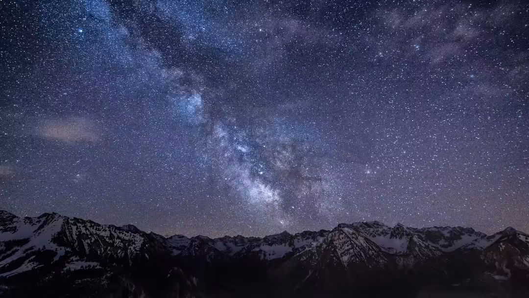 Milky Way galaxy stretching across night sky above snow-capped mountain peaks in deep blue and purple darkness with countless