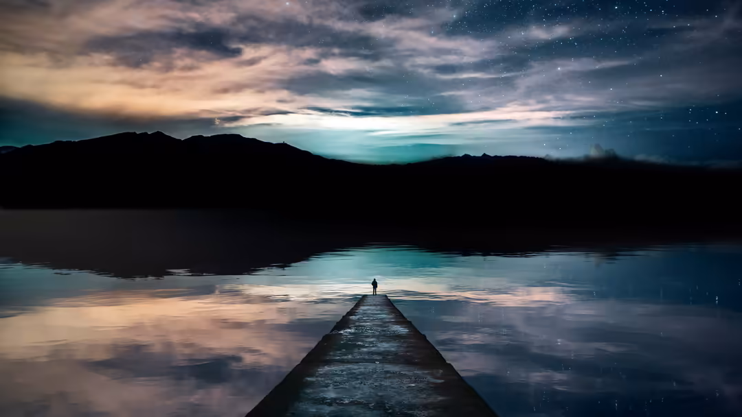 A solitary figure stands on a wooden pier extending into a calm lake, gazing at a starlit sky and distant mountains silhouett