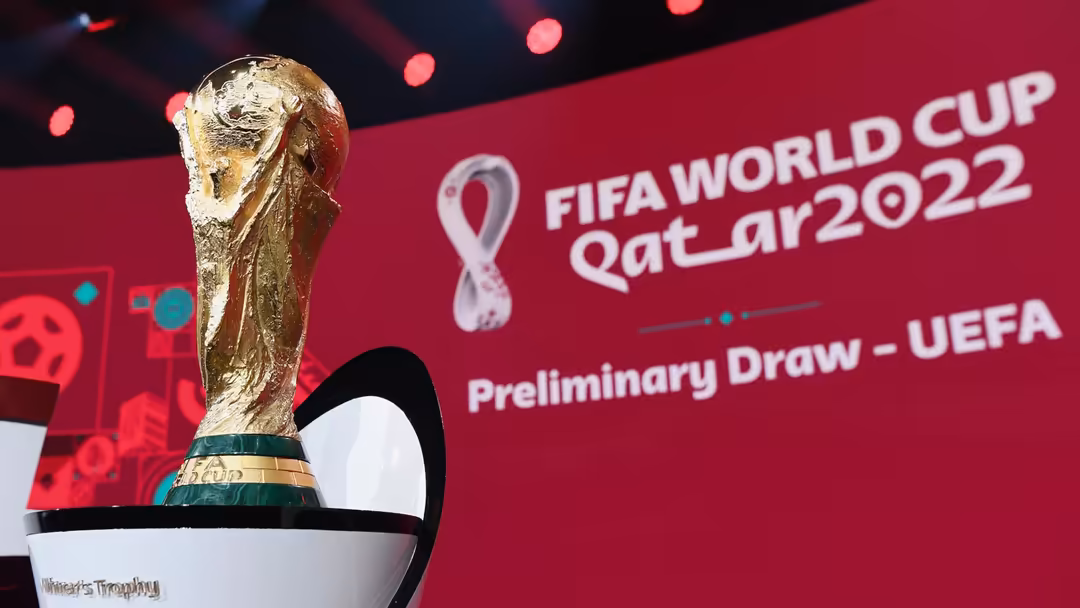 Golden FIFA World Cup trophy displayed on stage with Qatar 2022 branding and red background with bokeh lighting effects.