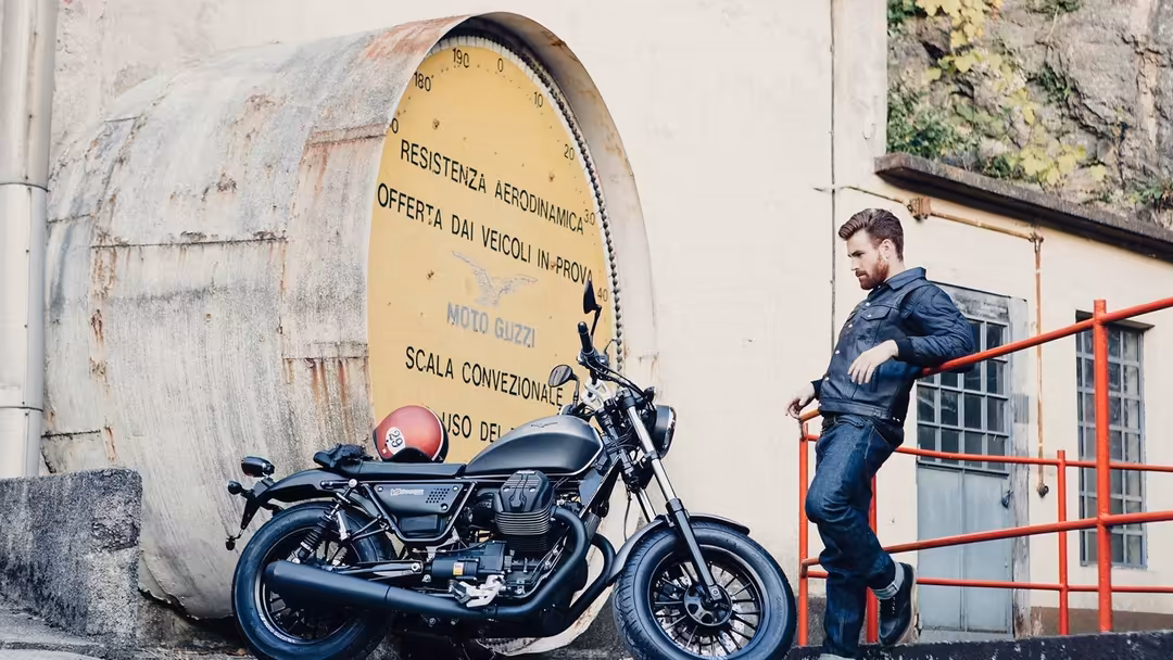 Man in denim jacket leaning against red railing beside classic motorcycle parked in front of weathered industrial wall with y