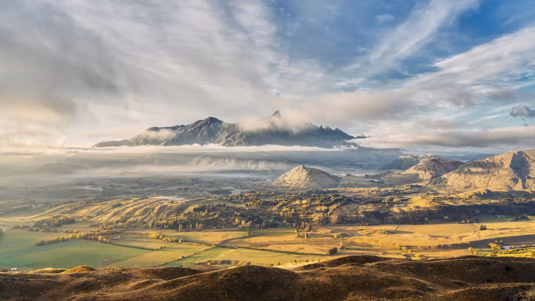Aerial view of a misty mountain peak rising above a golden valley with rolling hills and scattered buildings under a dramatic