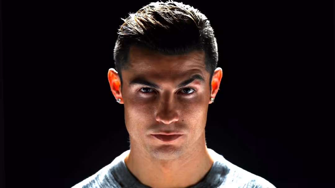 Cristiano Ronaldo intense portrait with blonde-highlighted dark hair against black background, wearing light gray shirt with