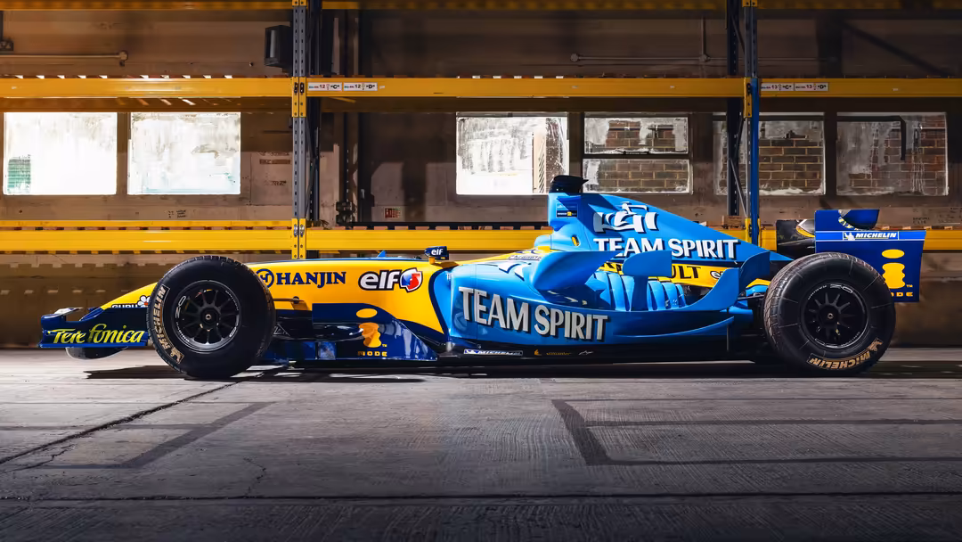 Blue and yellow F1 formula one race car with Team Spirit livery parked against industrial warehouse backdrop with yellow safe