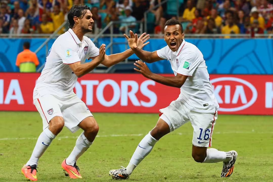 Soccer players in white uniforms celebrating with raised hands and excited expressions during a World Cup match with crowd in